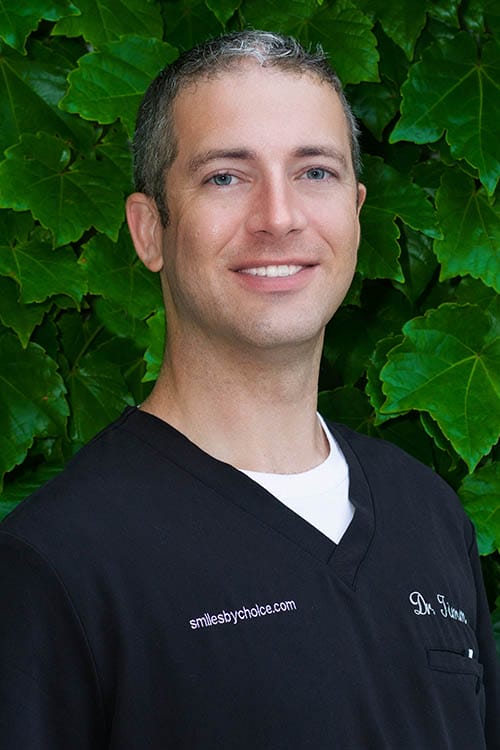 Dr. Adam Timm smiling in black scrubs, against a green leafy background