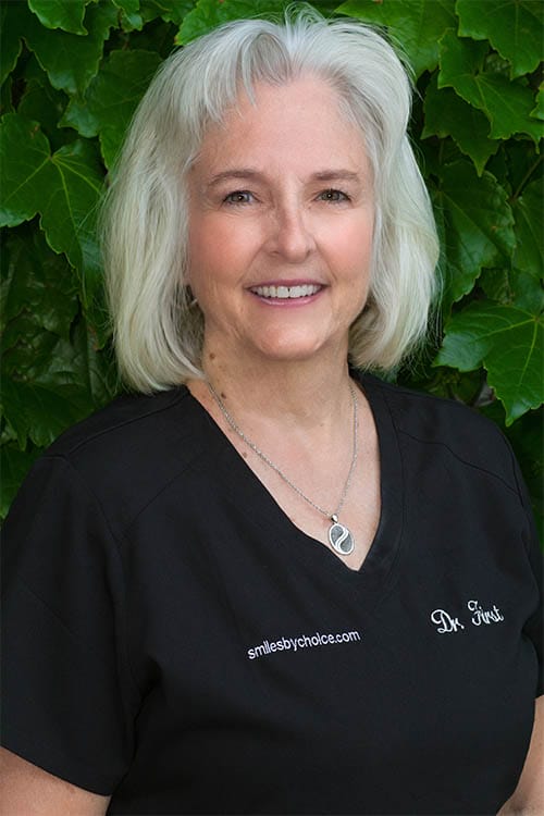 Dr. Louise First wearing black scrubs, smiling in front of green leafy background