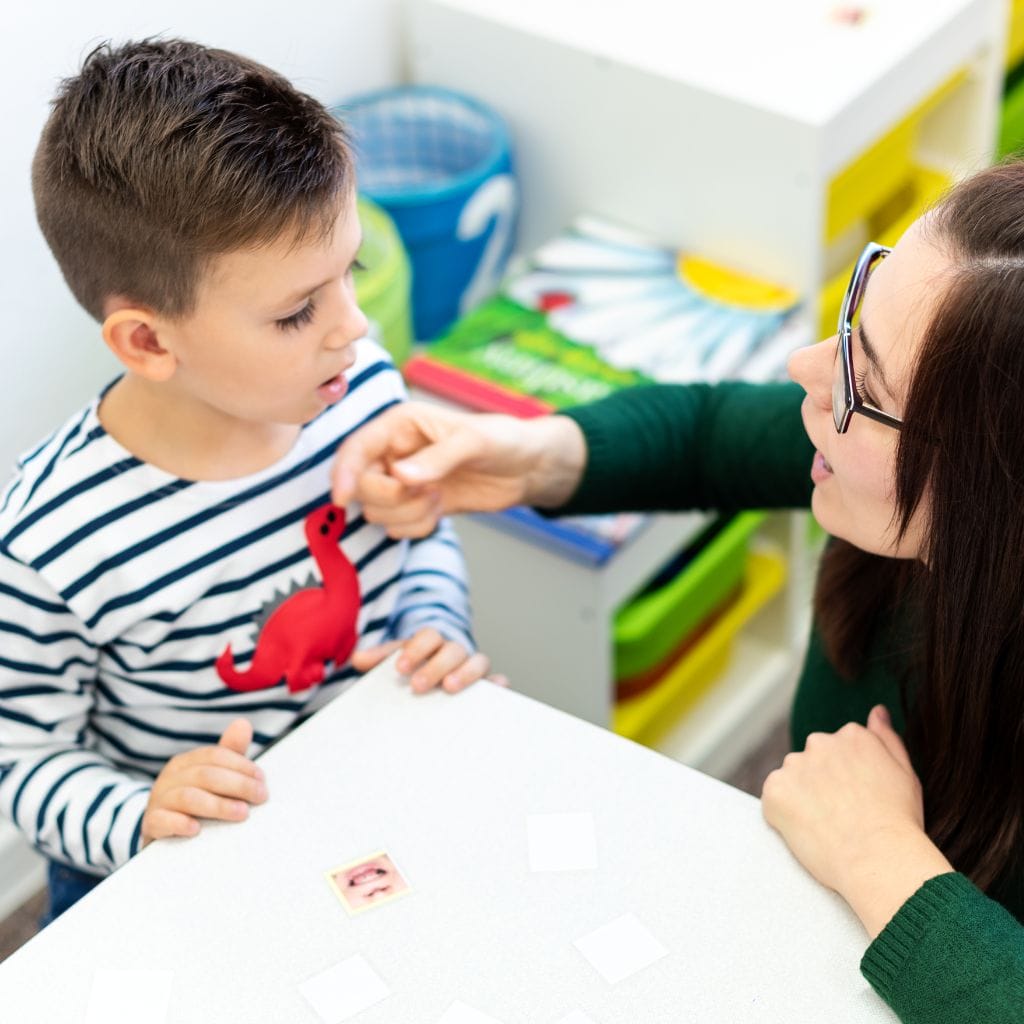 A woman with glasses and dark hair leans toward a young boy wearing a striped shirt with a red dinosaur. They are sitting at a white table where the woman points toward the boy's face while he mimics her mouth movements. A small picture of a mouth and several blank cards are on the table between them.