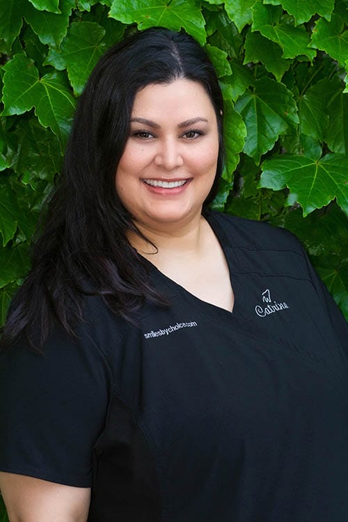 Catrina, CDA & EFDA in a black scrubs, smiling in front of a green leafy background