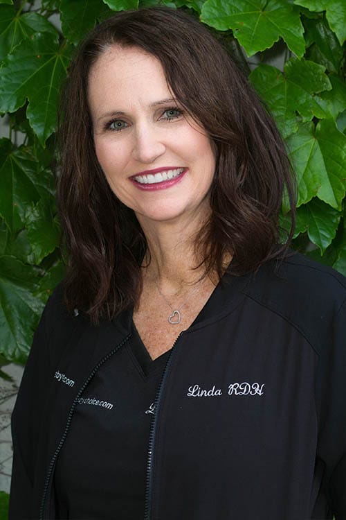 Linda, RDH wearing black scrubs, smiling in front of leafy plants
