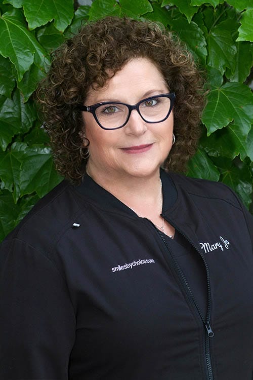 Mary Jo, CDA, with glasses wearing black scrubs in front of green leaves
