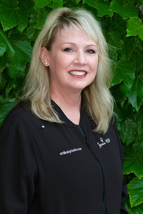 Jenise, RDH in a black scrubs, smiling in front of leafy greenery
