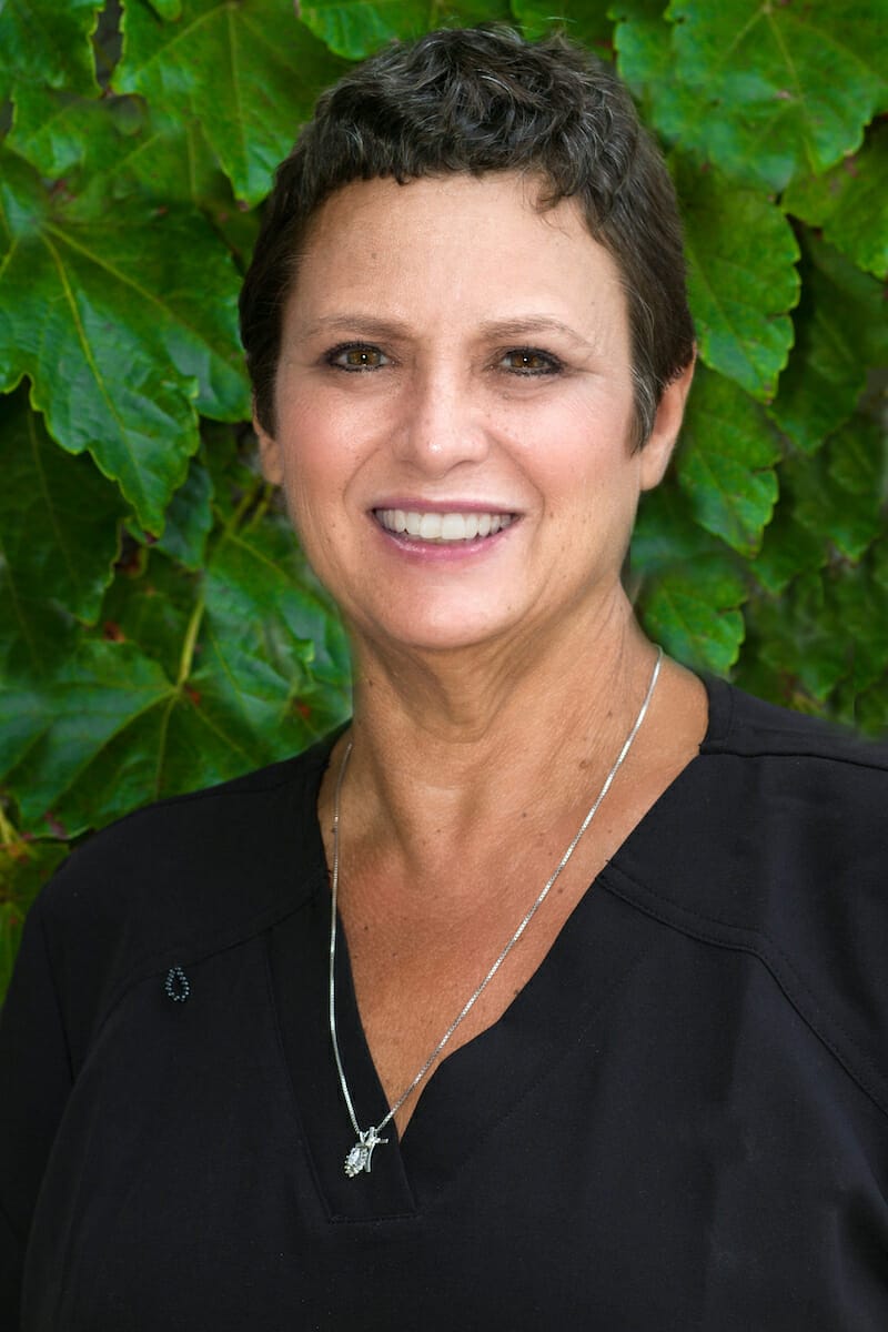 Justine, CDA, smiling in black scrubs and necklace, standing in front of leafy greenery