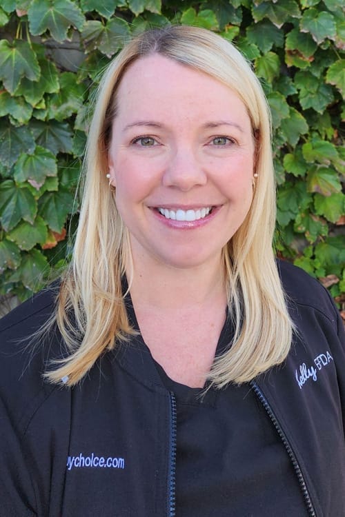 Kelly, CDA & EFDA in a black scrubs, smiling in front of a leafy wall